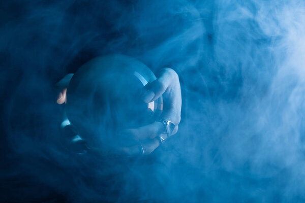 Обрезанный вид женских рук с хрустальным шаром и дымом вокруг на темно-синем фоне
