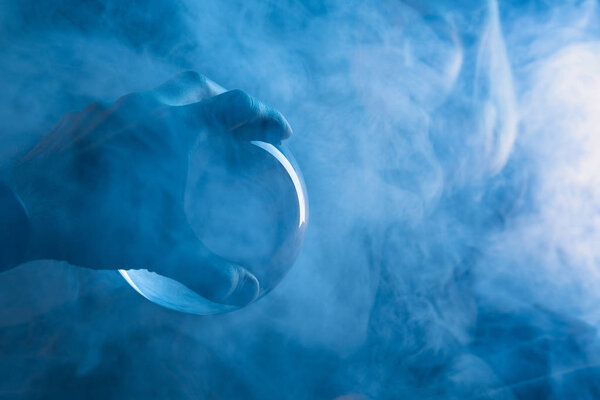 Cropped view of male hand with crystal ball and smoke around on dark blue