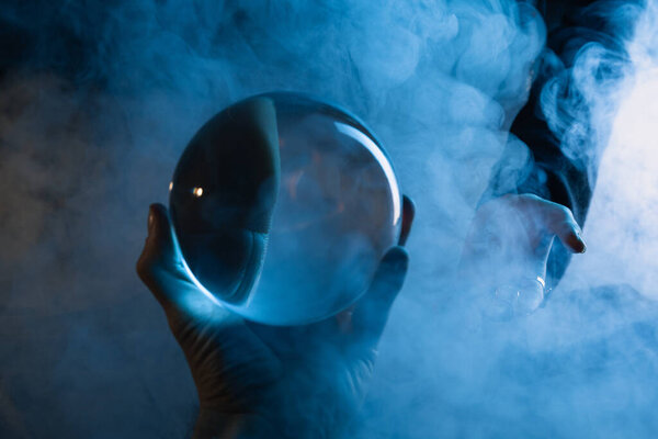 Cropped view of male hand with crystal ball and female hand with smoke around on dark blue