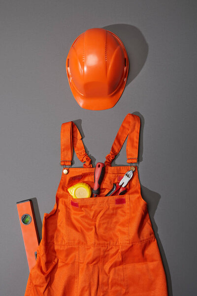 top view of orange helmet and overalls with measuring tape, screwdriver and pliers near spirit level on grey background