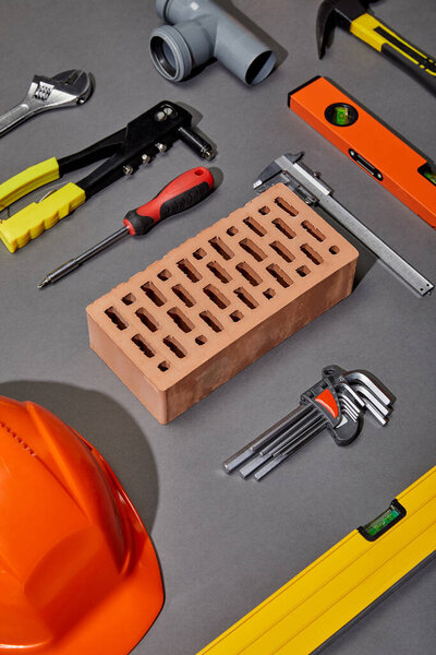 flat lay with orange helmet, brick, rivet gun, hammer, screwdriver, calipers, spirit level, angle keys and pipe connector on grey background