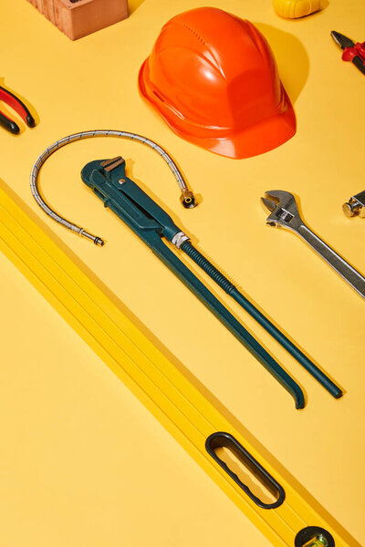 high angle view of calipers, helmet, monkey wrench, spirit level, brick, pliers and plumbing hose on yellow background