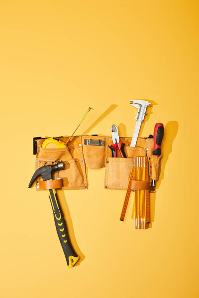 top view of tool belt with hammer, measuring tape, pliers, calipers, screwdriver and folding ruler on yellow background