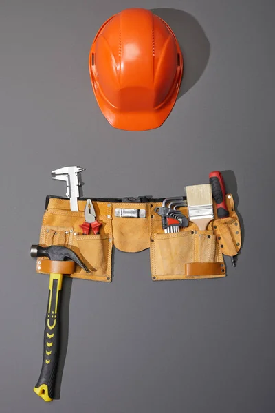 top view of helmet, tool belt with hammer, brush, pliers, calipers, angle keys and screwdriver on grey background