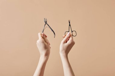 Cropped view of woman holding cuticle nipper and nail scissors isolated on beige clipart