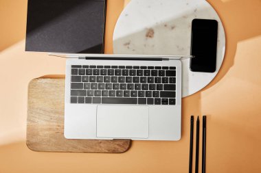 Tahta ve mermer tahtalardaki dizüstü bilgisayarın üst görüntüsü, akıllı telefon, boya fırçaları ve bej arka plandaki not defterleri