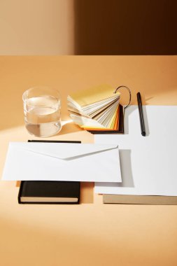 Kağıt yaprakları, renk örnekleri ve bej yüzey üzerindeki bir bardak su yakınındaki siyah defter üzerindeki zarf