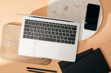 Tahta ve mermer tahtalardaki dizüstü bilgisayarın üst görüntüsü, akıllı telefon, not defterleri ve bej arka plandaki kalemler