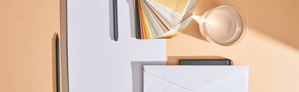 panoramic shot of glass of water, colors samples, sheets of paper, pens, envelope and notebook on beige background