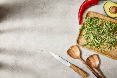 Mikro yeşillikli kesim tahtası, avokado yarısı, kırmızı biber, spatula ve gri arka planda bıçak.