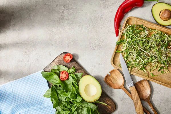 Top view of cutting boards with basil leaves, microgreens, cherry tomato and avocado halves with chili pepper, spatulas and knife on grey