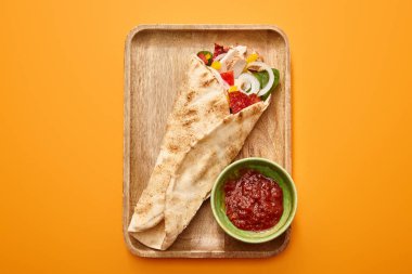 top view of fresh burrito with chicken and vegetables on board near chili sauce on orange background clipart