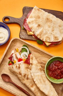 top view of fresh sandwich with salami in pita and burrito with chicken on boards near sauces on orange background clipart