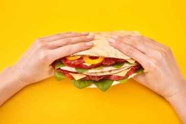 cropped view of woman holding fresh sandwich with salami, pita, vegetables and cheese on yellow background clipart