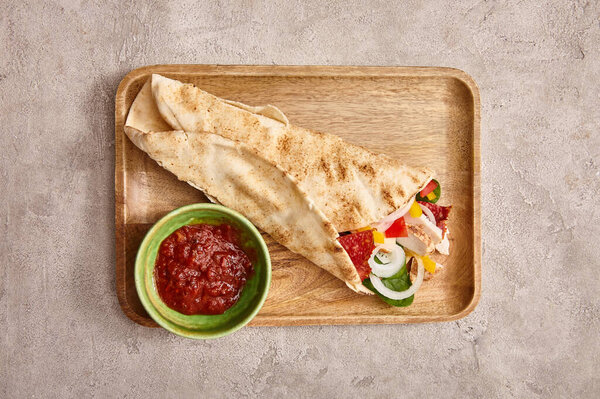 top view of fresh burrito with chicken and vegetables on board near chili sauce on concrete grey background