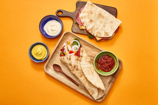 top view of fresh sandwich with salami in pita and burrito with chicken on boards near sauces on orange background