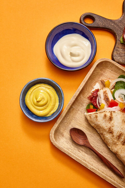 top view of fresh burrito with chicken and vegetables on board near chili sauce on orange background