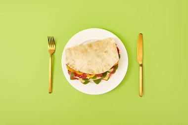 Altın çatalın yanında servis edilen tabakta salam, pide, sebze ve peynir ve yeşil arka planda bıçak ile taze sandviçin üst görüntüsü.