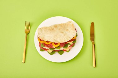fresh sandwich with salami, pita, vegetables and cheese served on plate near golden fork and knife on green background clipart
