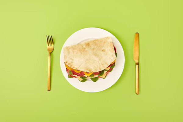 top view of fresh sandwich with salami, pita, vegetables and cheese served on plate near golden fork and knife on green background