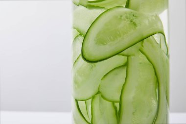 Close up view of glass bottle filled with liquid and sliced cucumbers on grey and white background clipart