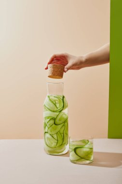Cropped view of female hand holding cork under bottle filled with water and sliced cucumbers on table on beige and green background clipart