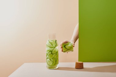 Cropped view of female hand holding glass filled with water and sliced cucumbers on beige and green background clipart