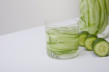 Kırpılmış şişe ve cam ile taze detoks içeceği ve gri üzerine ayrılmış dilimlenmiş salatalıkların yakın görüntüsü.