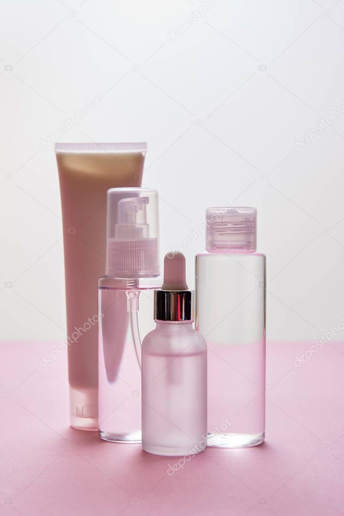 Set of cosmetic cream tube, cosmetic bottle, hand sanitizer spray and serum bottle with liquid on pink and grey background