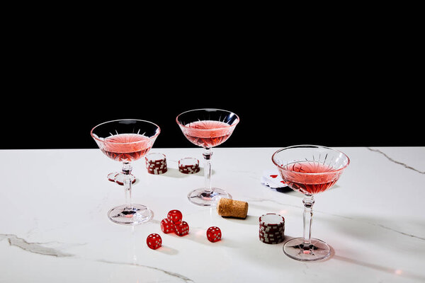 High angle view of glasses of cocktail, dice, cork, casino chips and pack of cards on white surface isolated on black