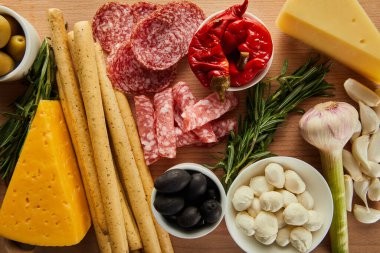 Tahta arka planda ekmek çubukları, salam dilimleri, peynir ve antipasto malzemelerinin üst görüntüsü