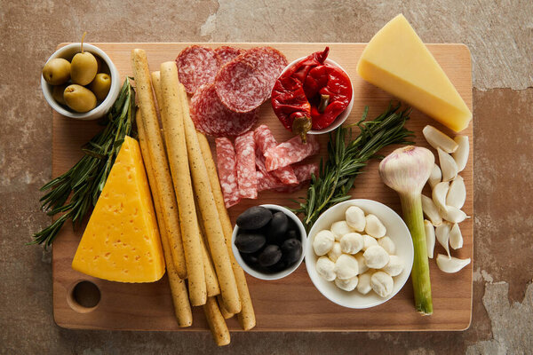 Top view of board with breadsticks, cheese, salami slices and antipasto ingredients on brown