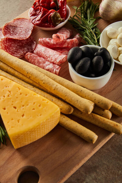 High angle view of cheese, breadsticks, salami slices, rosemary and bowls with antipasto ingredients on board on brown