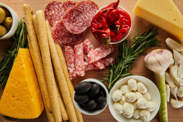 Top view of breadsticks, salami slices, cheese and antipasto ingredients on wooden background
