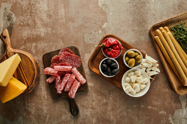 Top view of cheese, salami slices and antipasto ingredients on boards on brown background