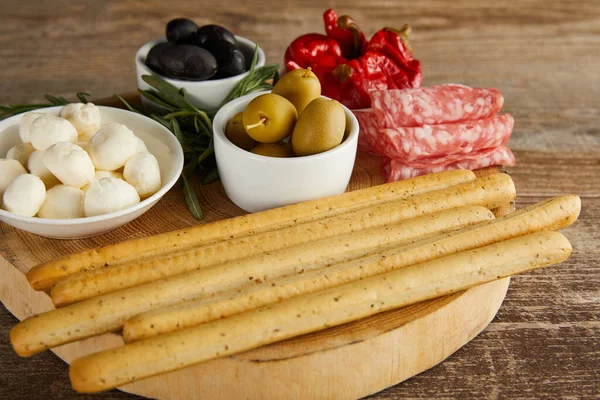 Round board with breadsticks, salami slices, greenery and bowls with antipasto ingredients on wooden background — Stock Image Board Breadsticks Salami Slices Greenery Bowls Antipasto Ingredients Wooden Background Stock Picture