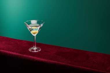 high angle view of cocktail glass with vermouth and whole olive on toothpick on velour surface on green background clipart