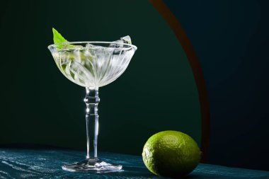 Buzlu kokteyl camının düşük açılı görüntüsü, nane yaprağı ve geometrik arkaplanda mavi ahşap yüzey üzerine tam limon.