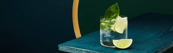 Glass with refreshing drink with lime slice, ice and mint leaf on wooden surface on geometric blue and green background