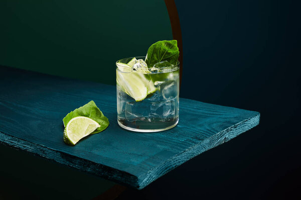 old fashioned glass with fresh drink, mint leaf and lime slice on blue wooden surface on green and blue geometric background