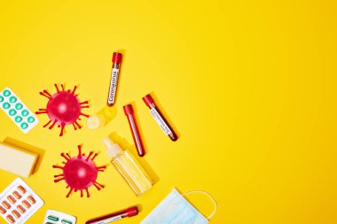 Tıbbi maskenin yanında koronavirüs testi harfi olan test tüplerinin üst görüntüsü, haplı su toplamaları, antibakteriyel sıvıyla şişeler ve sarı üzerine sabun çubuğu. 