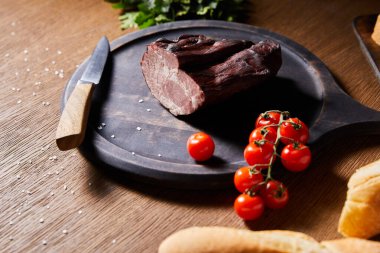 selective focus of tasty ham on board near parsley, cherry tomatoes and baguette with scattered salt and knife on wooden table clipart