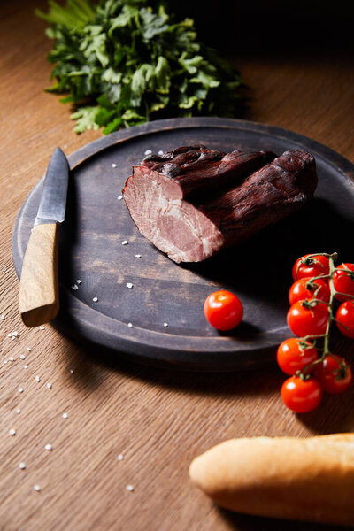 selective focus of tasty ham on board near parsley, cherry tomatoes and baguette with scattered salt and knife on wooden table