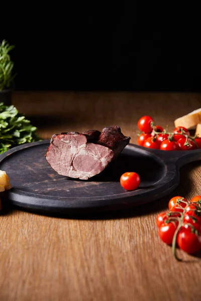 Selective focus of tasty ham on board near parsley, cherry tomatoes and baguette on wooden table isolated on black — Stock Image Selective Focus Tasty Ham Board Parsley Cherry Tomatoes Baguette Wooden Stock Picture