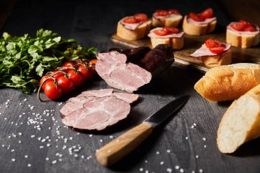 selective focus of tasty ham sliced ham, cherry tomatoes, parsley, salt, knife and baguette on wooden grey table with canape clipart