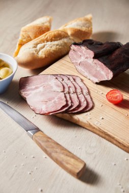 selective focus of tasty ham on cutting board with knife, cherry tomato, olives and baguette on wooden table clipart