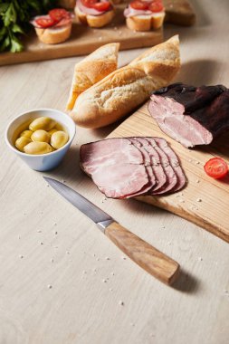 selective focus of tasty ham on cutting board with knife, olives and baguette on wooden table clipart