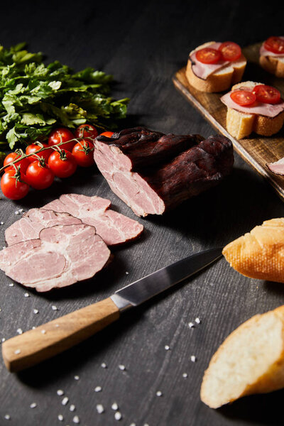 selective focus of tasty ham sliced ham, cherry tomatoes, parsley, salt, knife and baguette on wooden grey table with canape