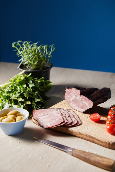 selective focus of tasty ham on cutting board with knife, parsley, cherry tomatoes, olives on wooden table on blue background