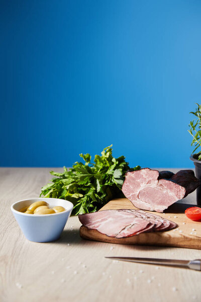 selective focus of tasty ham on cutting board with knife, parsley, olives on wooden table on blue background